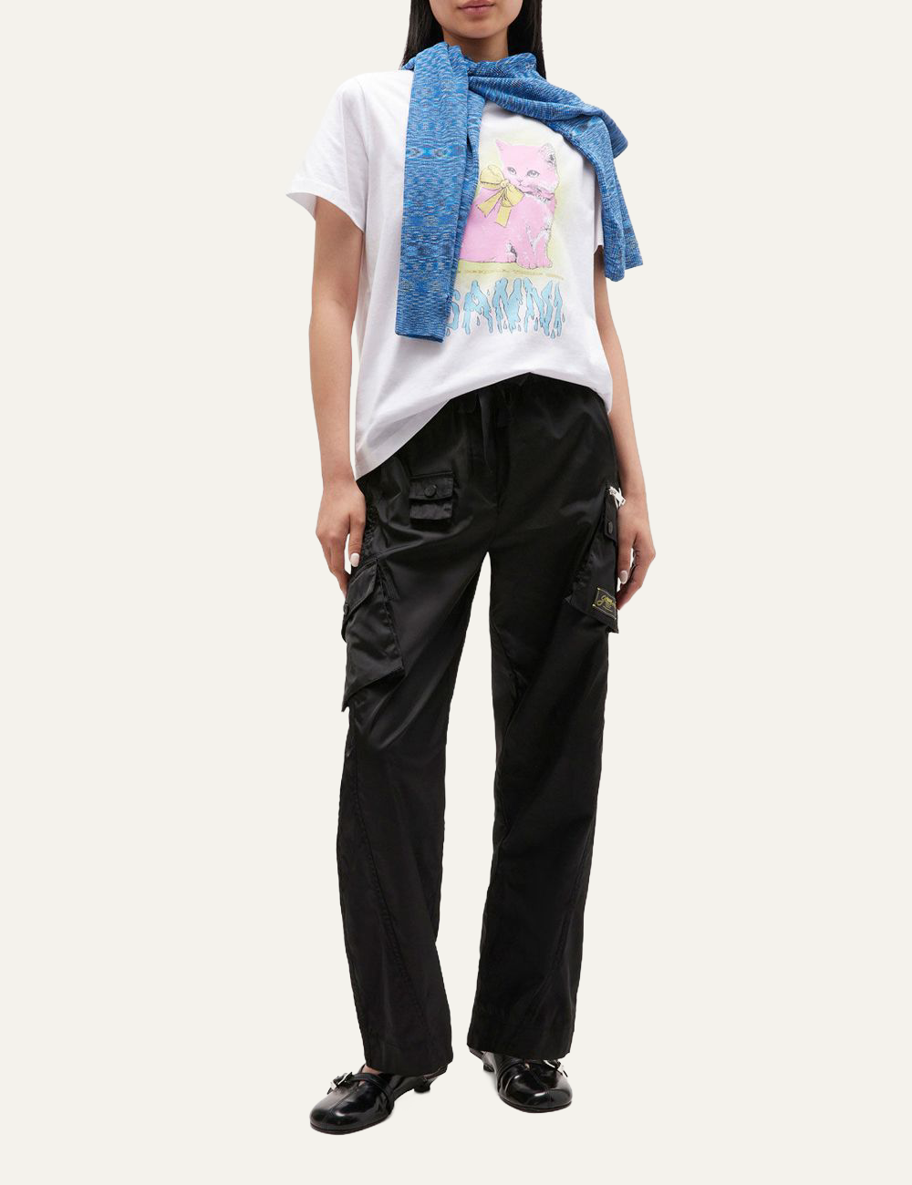 Full-body view of model in white Ganni T-shirt with pink kitten print, black cargo pants, and blue knitwear draped over shoulders.