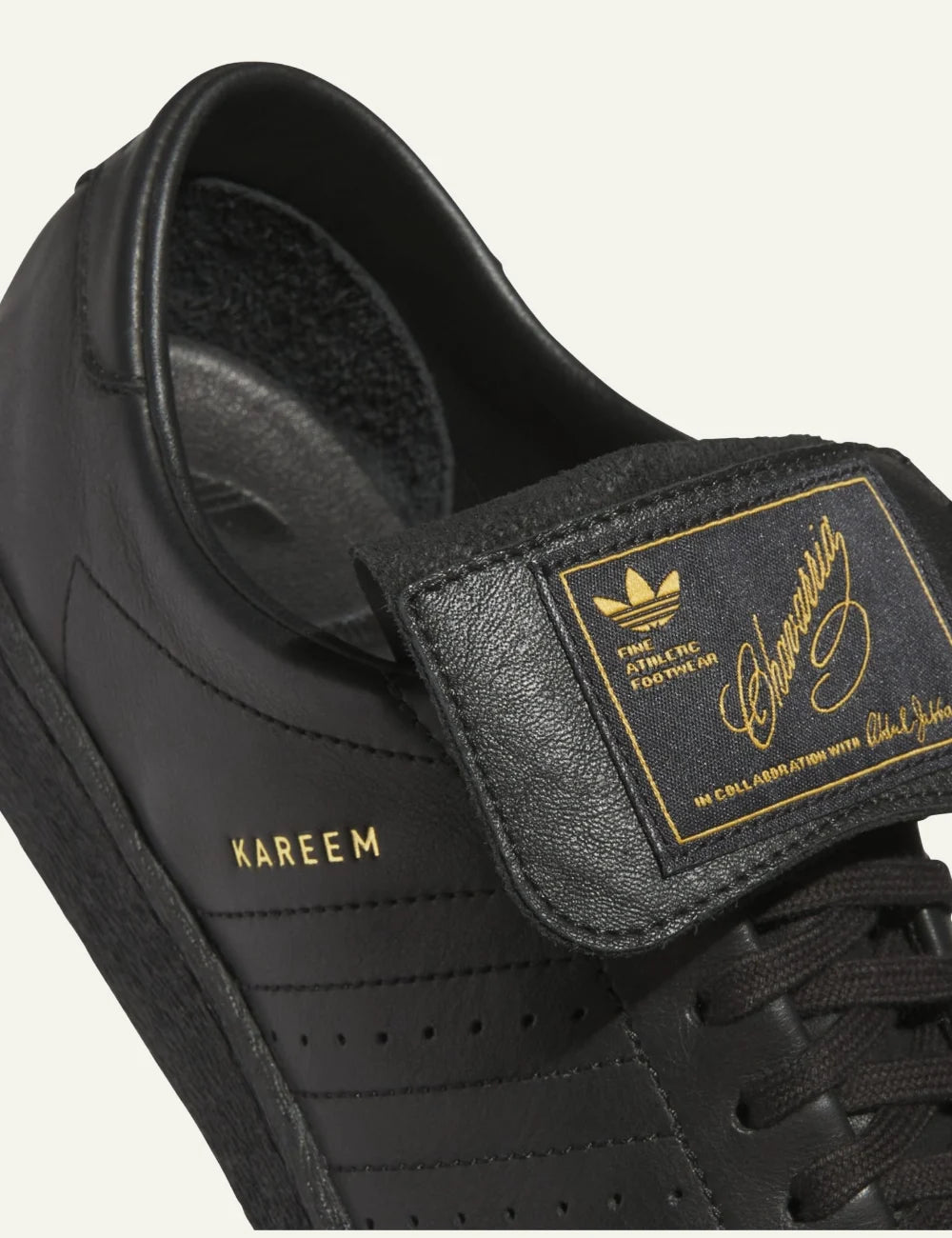Detail close-up: Gold signature Kareem Abdul-Jabbar branding on folded tongue σε μαύρο leather Adidas sneaker