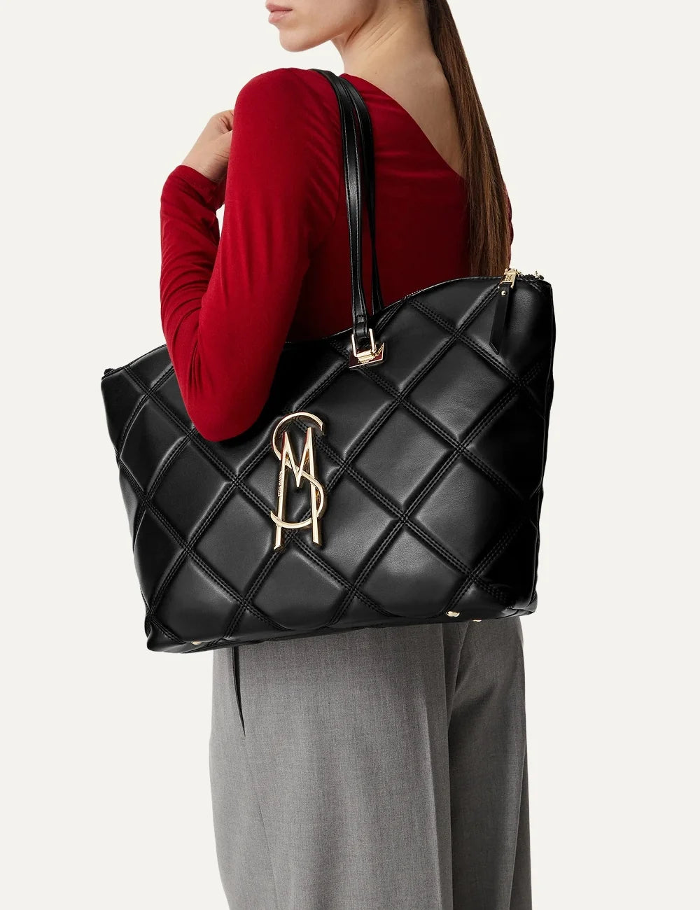 Steve Madden BVUSE women's black quilted faux leather tote bag with large diamond stitching, gold tone metal logo, two long shoulder handles and top zipper closure,  on model